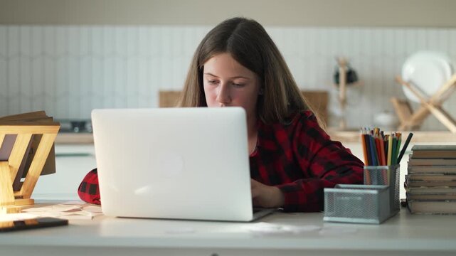 Focused teenage girl studying online using silver laptop. Young student sits at bright desk doing school homework. Diligent girl types on laptop keyboard. Tough homework requires deep studying.