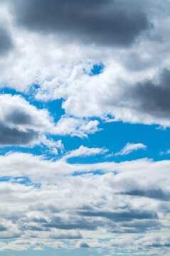 Picturesque blue sky texture, vertical vibrant cloudscape with a mix of fluffy white cumulus clouds, darker overcast storm clouds, and windows of bright blue sky