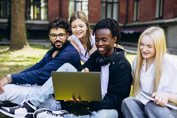 Group of happy diverse university students sitting on grass, looking at a laptop and smiling...