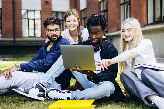 Diverse group of university students using a laptop for research and teamwork on campus grass, fostering learning and friendship
