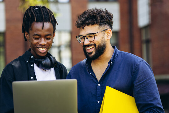 Two smiling male students of different ethnicities collaborating on a laptop, standing outdoors on a university campus