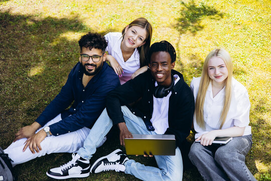 Diverse college students smiling, sitting on green grass using a laptop and notebook, enjoying friendship outdoors