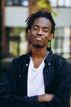 Young black man with dreadlocks smiling, standing with arms crossed, portraying confidence and happiness outdoor