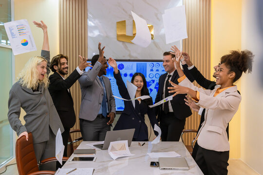 ​A multi-ethnic professional team celebrating a project completion by tossing papers in the air at a modern boardroom, expressing joy, teamwork, and corporate achievement.