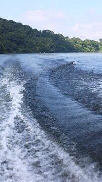 Powerful boat wake flowing toward dense tropical shoreline with Bali nature, pale blue sky, soft clouds and calm water reflections over open water in low aft coastal view
