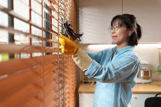 Woman dusts and wipes wooden window blinds with a microfiber cloth, promoting hygiene, cleanliness, and efficient home maintenance in a bright modern interior.
