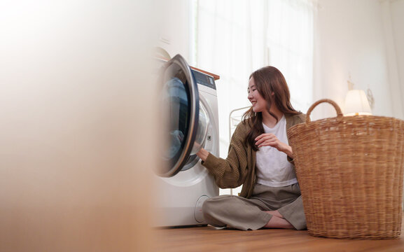 Smiling Asian woman loading clothes into a washing machine with a laundry basket, reflecting modern home care, laundry routine, cleanliness.