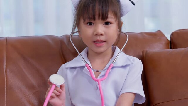 Asian girl child dressed as nurse with stethoscope sitting on couch, representing education, career aspiration, healthcare concept, and childhood imagination in cheerful indoor environment