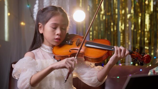 Asian girl child playing violin indoors, representing education, training, discipline, and childhood development in cheerful environment with warm lighting, decorative bokeh, and positive atmosphere