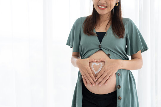 Pregnant woman showing belly with cream heart symbol, highlighting maternity care, love, and wellness in cozy indoor environment with natural daylight, lifestyle branding, and positive family concept