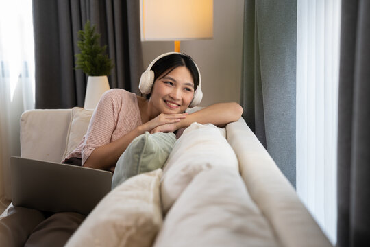 Asian woman resting with headphones and laptop in cozy living room, highlighting leisure, digital entertainment, and home lifestyle with warm lighting, cushions, and comfortable domestic environment
