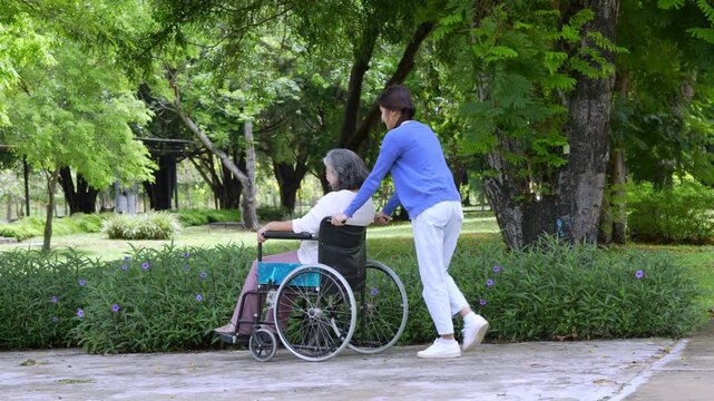 Caregiver daughter helping elderly mother sit in wheelchair at park, representing family bonding, senior care, wellness, and companionship in cheerful outdoor environment with trees, and daylight.