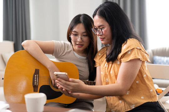 Two friends practicing guitar together at home, using a smartphone for chords or music notes. Promotes friendship, creativity, and modern lifestyle through collaboration, technology