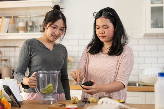 Asian women enjoying cooking practice by preparing smoothie with fruits in bright modern kitchen, emphasizing wellness, friendship, and healthy lifestyle through teamwork, blender use, and cheerful