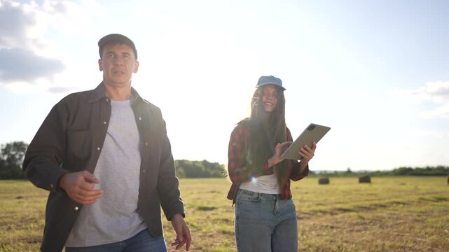 Two people standing in field with tablet. Woman and man working in a field with hay. Farmers studying working lifestyle in fields with haystacks. A couple of individuals in a field holding a tablet.