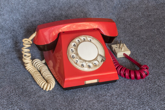 A vintage landline telephone in a red case with a classic rotary dial and a white, coiled, stretchable handset cord. A classic four-pin telephone plug on a red cord is visible on the right.