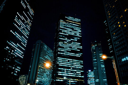 夜の新宿の高層ビル群を見上げるアングルで撮影した写真。窓から漏れる無数の照明が都会の夜景を彩り、洗練されたビジネス街の冷徹さと近未来的な雰囲気が漂う様子を捉えている。