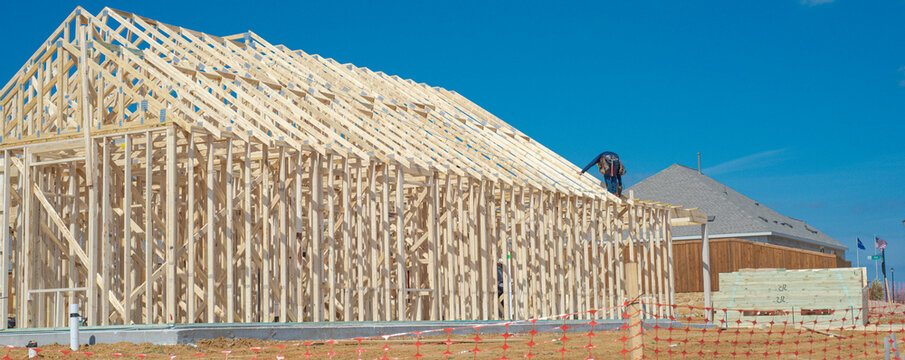 Laborer maneuvers across high rafters atop Dressage Ln property, showcasing precise vertical development. Finished house contrasts with raw building materials beneath sunlight. Aubrey, TX