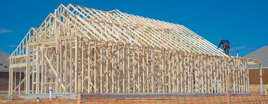Workman maneuvers across peak roof rafters overlooking residential plot near Dressage Ln. Intricate wood framed geometry marks vital building phase before exterior materials arrive. Aubrey, TX