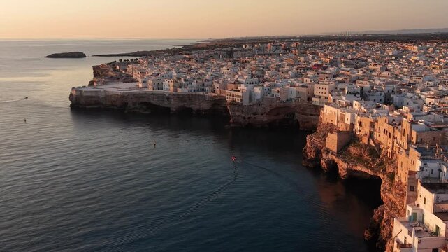 Aerial view of polignano a mare, a picturesque town built on cliffs overlooking the adriatic sea, with a small boat sailing into a sea cave during a warm sunset