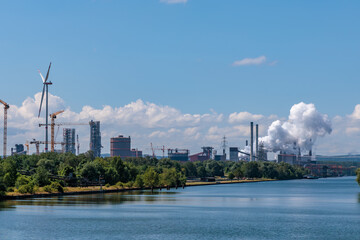 Obraz premium Panoramic view of an industrial plant with wind turbine, smoke stacks and cranes along a river waterway.