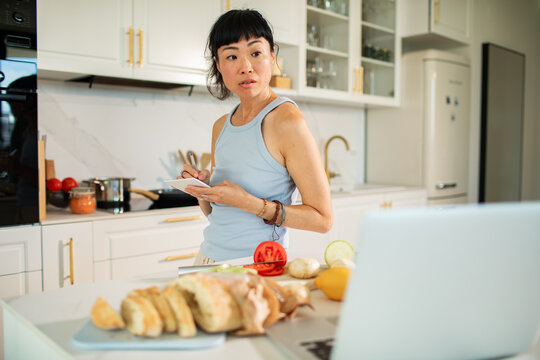 Woman taking recipe notes while cooking with laptop in modern kitchen