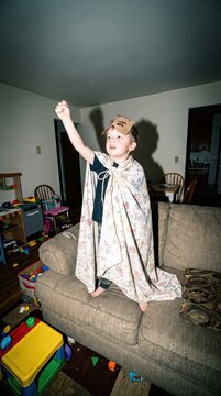 little caucasian boy in makeshift superhero costume with blanket cape and cardboard mask standing on couch with raised arm. pretend play, childhood imagination. messy living room background.