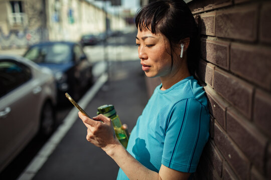 Woman with earbuds checking phone on city sidewalk after workout