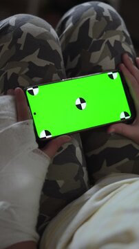 Hands of caucasian child with cast holding smartphone with green screen. Boy injury and use of gadgets for viewing.