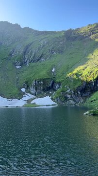 Vertical shot of a slow pan over Balea Lake in warm morning light showing Cabana Balea Lac chalet and Fagaras mountain peaks along Transfagarasan road, Romania. Scenic summer travel destination.