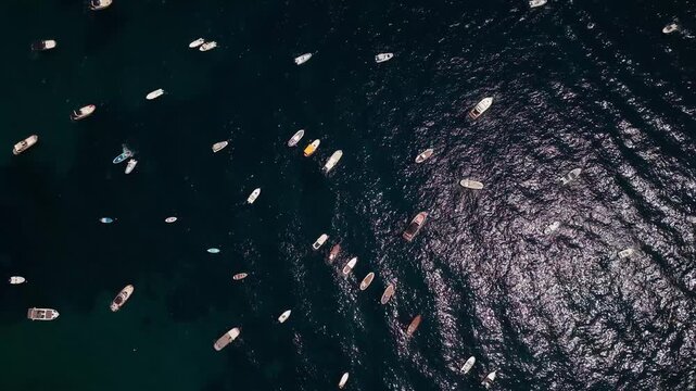 Aerial top-down view of many boats, yachts, and ships anchored in a dark ocean with sun glistening on the rippling water, revealing a serene maritime landscape