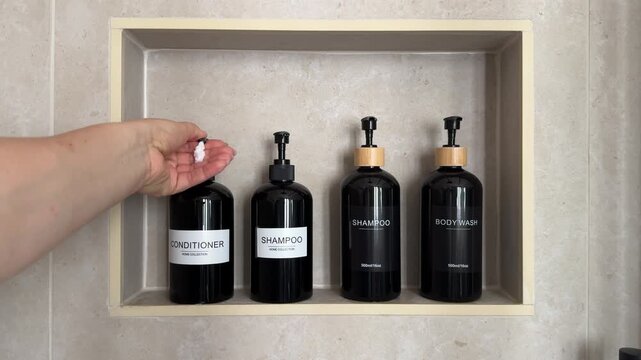 Hand dispensing conditioner from a refillable pump bottle in a built in shower niche with shampoo and body wash containers. Bathroom organization, personal care and sustainable refill concept for home