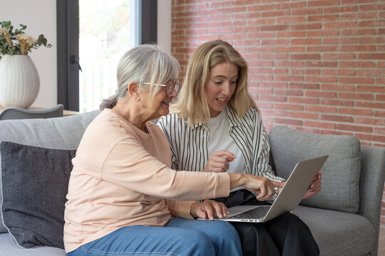 Young woman teaching mature adult senior lady how to use a laptop, two women sitting on a sofa at home, sharing online content, generational learning