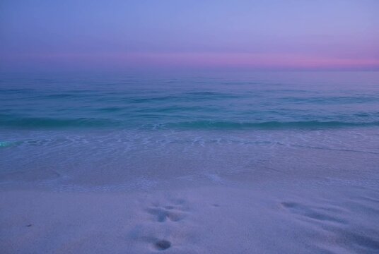 Tranquil Beach Twilight: The gentle ebb and flow of turquoise waves kiss the shore of a serene sandy beach, leaving fleeting footprints under a soft.