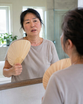Midlife Asian woman fanning herself in a bathroom mirror during a hot flash, showing facial flushing, discomfort, menopause symptoms, and self-awareness.