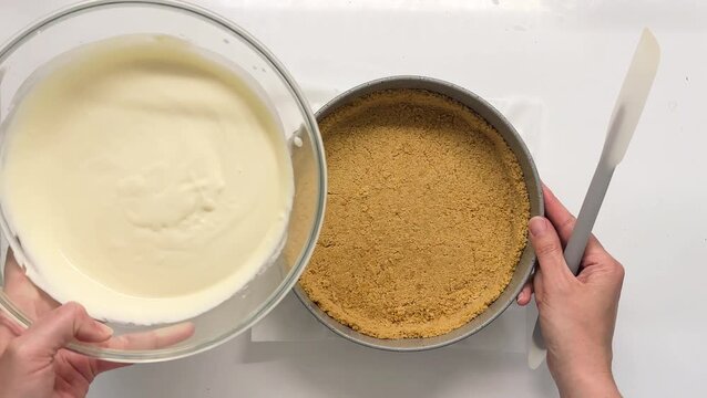 Overhead view of hands pouring smooth cream cheese batter into a springform pan with graham cracker crust and spreading it with a silicone spatula for a homemade cheesecake