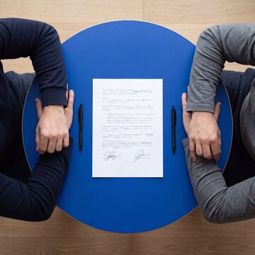 Two business partners with folded arms sitting at round blue table with signed contract document and pens in top view, negotiation and agreement concept.