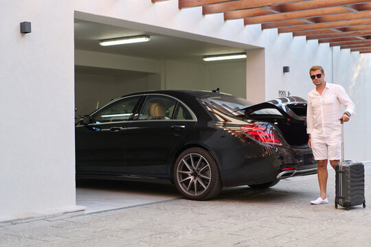 Man With Suitcase Standing by Luxury Black Car in Garage Near Pool at Resort Arrival