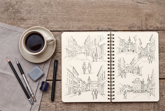 Open sketchbook with urban city drawings beside coffee cup and pencils on rustic wooden table, top view of sketching hobby and creative analog leisure.