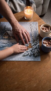 Parent and child hands assembling winter jigsaw puzzle on wooden table with candle and cocoa mugs capturing cozy family leisure during holiday season.
