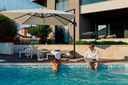 Man Working On Laptop By Pool While Child Plays At Modern Home Patio Under Umbrella
