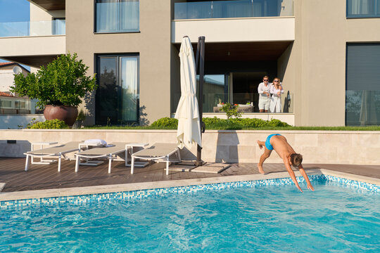 Young Boy Diving Into Private Pool While Parents Watch From Modern Villa Balcony On Sunny Morning