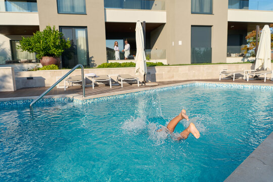 Couple Watching Child Splashing Into Hotel Pool At Sunny Resort With Balconies And Sun Loungers