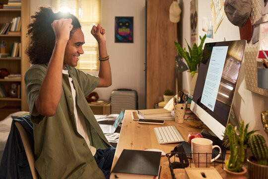 Young adult man celebrating at desktop computer while reading acceptance message in home workspace, expressing success and achievement. Ideal for education, remote work, career growth
