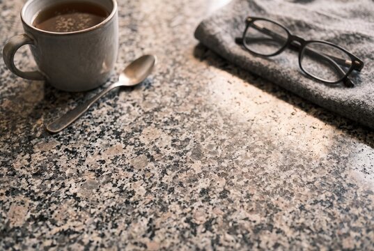 Coffee mug with vintage spoon and eyeglasses on folded knit sweater over speckled granite countertop in warm morning sunlight with copy space.