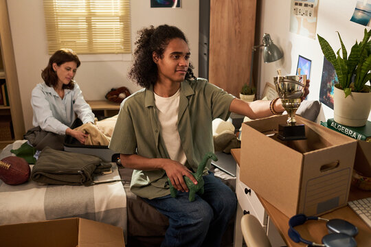 Teenage son packing belongings in bedroom while mother organizing suitcase, preparing for college move and family transition. Ideal for education, parenting, relocation marketing