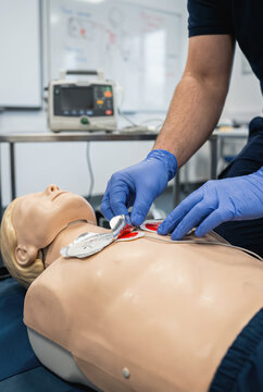 A closeup reveals gloved hands applying defibrillator pads on a mannequin in a medical training setting, demonstrating proper technique with nearby equipment visible in the background