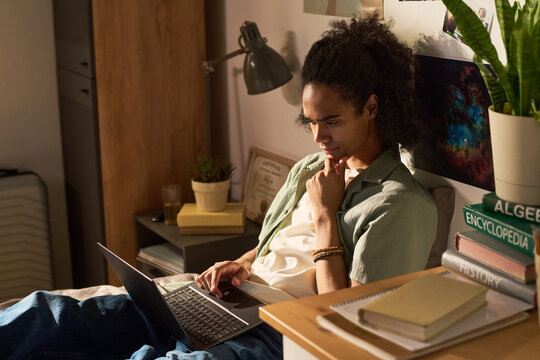 Young adult Black man studying on laptop in bedroom, researching coursework and thinking critically. Ideal for online education, remote learning, student productivity themes
