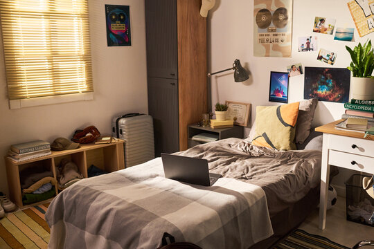 Wide angle shot of student bedroom featuring laptop on bed, study desk, books, suitcase, personal decor, suggesting college living, remote learning, youth lifestyle marketing
