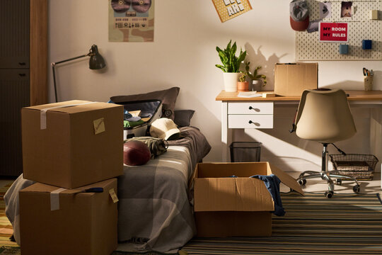 Wide shot of teenager bedroom showing cardboard boxes, packed suitcase, desk, bed, signaling move in or move out. Supporting relocation, student housing, dorm life, transition marketing